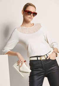 Witte top met lange mouwen en kanten inzetstuk, zwarte strakke broek, witte textuurrijke clutch en rode hoekige zonnebril. Riem met gouden accenten.