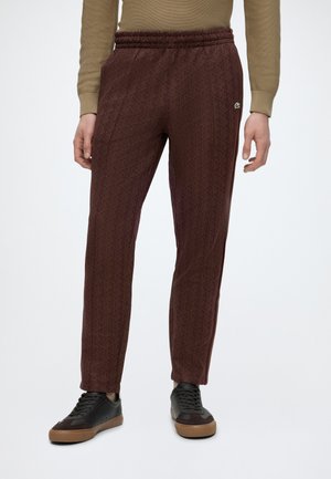Persona che indossa pantaloni marroni con motivo, sneaker nere e marroni e una maglia beige a maniche lunghe, in piedi contro uno sfondo semplice.