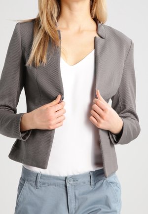 Kvinde iført en tekstureret grå blazer over en hvid top og lyseblå bukser, der justerer blazeren med begge hænder.