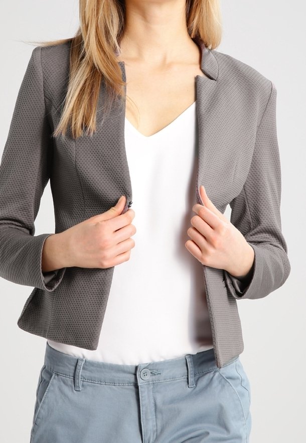 Femme portant un blazer gris texturé sur un haut blanc et un pantalon bleu clair, ajustant le blazer avec les deux mains.
