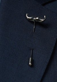 Silvrig mustasch-nål med spetsig baksida, fäst vid en strukturerad marinblå textil, med en distinkt böjd design och polerad yta.