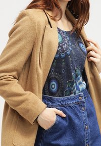 Brauner Wollmischblazer über einem dunkel gemusterten Oberteil mit blauen und grünen kreisförmigen Designs, kombiniert mit einem hochtaillierten Jeansrock.