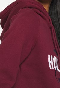 Felpa con cappuccio color marrone scuro con tessuto testurizzato, caratterizzata da un logo bianco e dettagli ricamati a contrasto. Il cappuccio è dotato di coulisse per la regolazione.