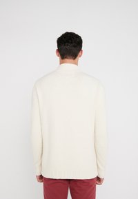 Suéter de punto color crema con un cuello alto, tejido texturizado y puños y dobladillo acanalados. Combinado con pantalones rojos.