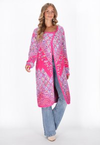 Roze en paarse lange cardigan met een fuzzy textuur, voorzien van geometrische patronen en een brede opening, gecombineerd met lichtblauwe jeans.