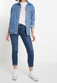 Veste en denim bleu clair, avec des poches poitrine et des manches retroussées, portée sur un t-shirt graphique rose. Jean skinny bleu foncé et baskets blanches.