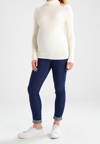 Białe sweter z golfem w prążki w połączeniu z ciemnoniebieskimi skinny jeansami i białymi sneakersami. Jeansy mają podwinięte nogawki, co nadaje swobodny wygląd.