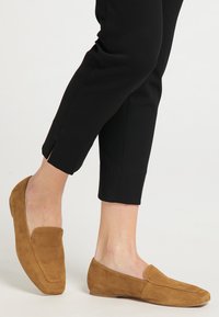 Mocassini din suede maro, cu toc plat, având o textură netedă și un design cu vârful rotund, asortați cu pantaloni negri eleganți.