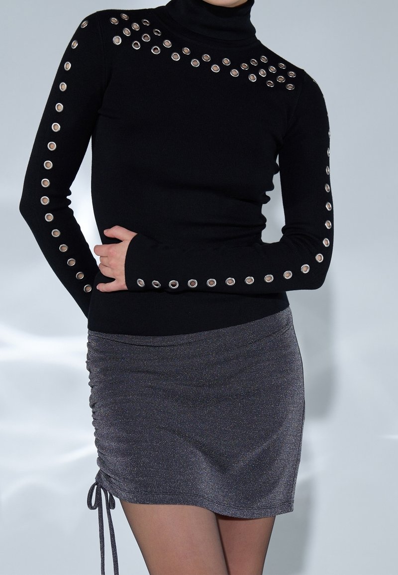 Schwarzer Rollkragensweater mit großen metallischen Ösen an Schultern und Ärmeln, kombiniert mit einem grauen schimmernden Minirock mit Kordelzügen.