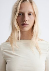 Beige long-sleeve top with a round neckline, featuring light texture and side ruching. The model has straight, light blonde hair.