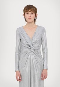 Ασημί φόρεμα με μακριά μανίκια, διαθέτει βαθύ V-λαιμόκομμα και στριφτό σχέδιο στη μέση, κατασκευασμένο από υλικό που λαμπυρίζει και αντανακλά το φως.