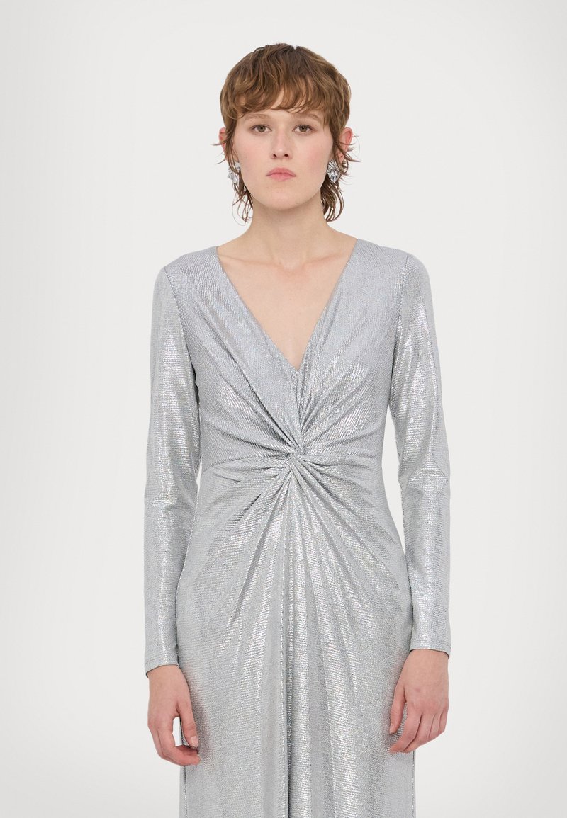 Ασημί φόρεμα με μακριά μανίκια, διαθέτει βαθύ V-λαιμόκομμα και στριφτό σχέδιο στη μέση, κατασκευασμένο από υλικό που λαμπυρίζει και αντανακλά το φως.
