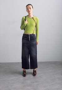 Maglietta verde a coste con maniche lunghe, dotata di una fascia diagonale marrone e dettagli con bottoni, abbinata a jeans neri a gamba larga con chiusura a bottone in argento. Scarpe marroni a punta quadrata.