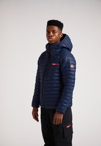 Mørkeblå puffer jakke med hætte, der har et rødt logo patch på brystet og et rundt logo på ærmet. Struktureret, quiltet design.