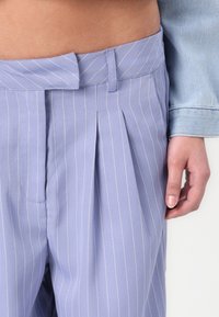 Ανοιχτό μωβ πλισέ παντελόνι με διακριτές λευκές ρίγες, που διαθέτει επίπεδη ζώνη και πλαϊνές τσέπες. Το υλικό φαίνεται λείο και ελαφρύ.