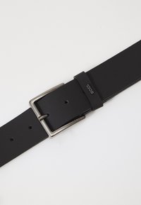 Zwarte leren riem met een rechthoekige zilveren metalen gesp, voorzien van een gestructureerde afwerking en meerdere aanpassingsgaten. Merken zijn in de band gestempeld.