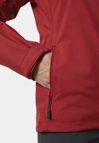 Rode outdoor jas met een gestructureerde afwerking, voorzien van een zijvak met ritssluiting en verstelbare mouwen. Materiaal lijkt duurzaam en licht van gewicht.
