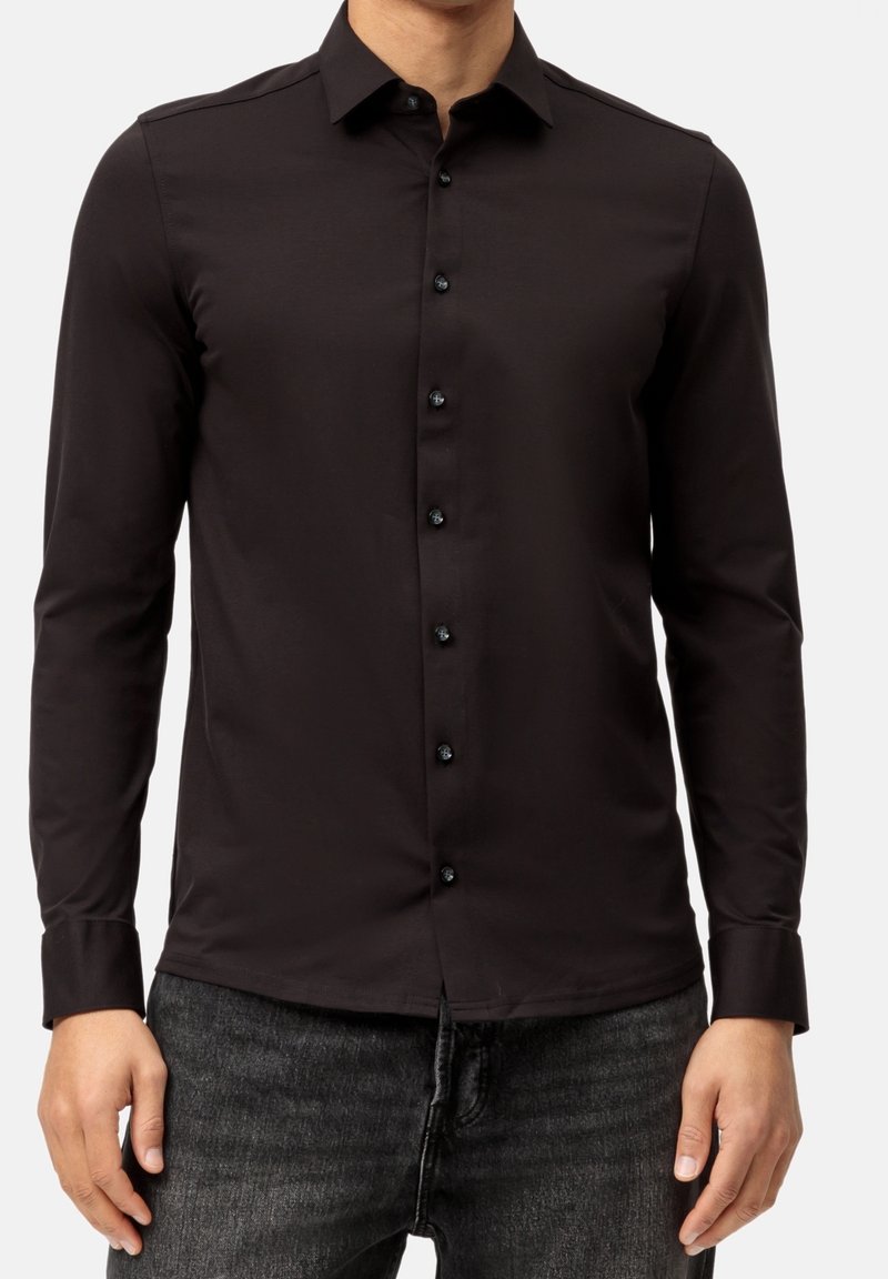 Homme portant une chemise noire ajustée à manches longues avec un col pointu et un jean gris foncé, sur un fond uni.