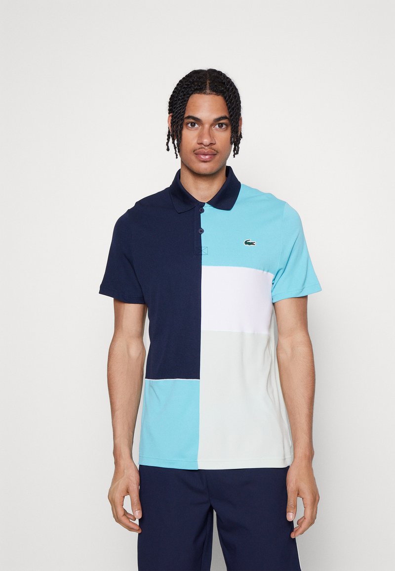 Polo shirt coloré en blocs de bleu marine, bleu clair et blanc. Manches courtes, patte de boutonnage, logo Lacoste sur la poitrine, en coton.