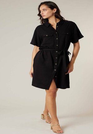 Femme portant une robe chemise noire ceinturée à manches courtes et des sandales à talons blocs ouverts dorées, debout devant un fond uni.