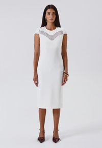 Donna che indossa un vestito midi bianco con dettagli in pizzo sul petto, tacchi bordeaux e tre braccialetti sul polso destro, in piedi contro uno sfondo semplice.
