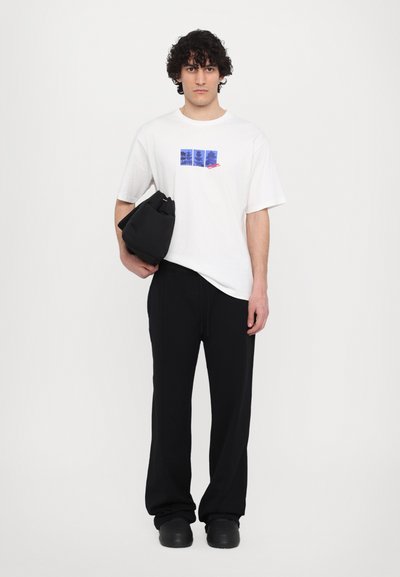 T-shirt blanc avec un design graphique bleu à l'avant, associé à un pantalon noir ample et tenant un sac noir. Texture lisse.