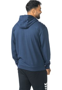Marineblauwe hoodie met een zachte textuur, geribbelde manchetten en een logodetail op de linkerschouder. Achteraanzicht toont een losse pasvorm.