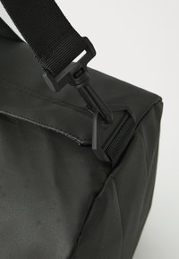 Sac en tissu noir avec une sangle robuste, doté d'un clip en plastic noir et de coutures. La texture est lisse avec une finition mate.