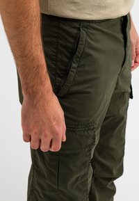 Olijfgroene cargobroek met zijrits zakken, textuurstof, elastische tailleband en gestikte accenten. Close-up weergave toont hand op de naad.