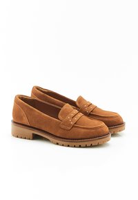 Mocassins en daim beige avec un bout rond, une sangle décorative avec des clous en métal, et une semelle en caoutchouc brun clair. Texture lisse sur l'ensemble.