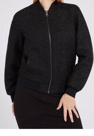 Frau trägt eine schwarze, strukturierte Bomberjacke mit Reißverschluss, gerippten Bündchen und Bund, kombiniert mit einem schwarzen Rock, Hand auf der Hüfte ruhend.