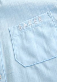 Svijetloplava tkanina ima suptilni teksturirani uzorak, a pravokutna šupljina prikazuje izvezeno ime "BAKER" u bijeloj boji.