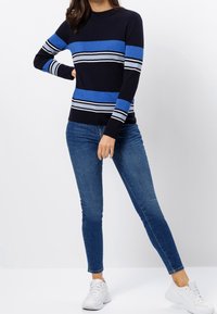 Woman wearing navy blue sweater with blue and white stripes, blue skinny jeans, and white sneakers, standing with one hand on hip.