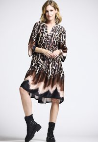 Braun- und schwarz geflecktes Kleid aus leichtem Stoff, mit gestuftem Design, V-Ausschnitt und Dreiviertelärmeln, kombiniert mit schwarzen Kampfstiefeln.