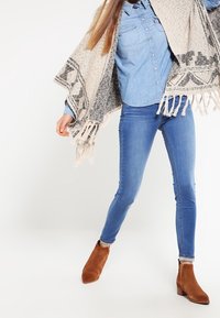 Poncho beige con patrones geométricos negros y bordes con flecos, llevado sobre una camisa de botones azul claro, combinado con jeans ajustados azules y botas de tobillo marrones.