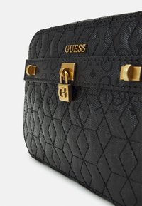 Svart Guess-väskan med en texturerad diamantmönster, detaljer i guld och låsöppning. Kompakt rektangulär form.