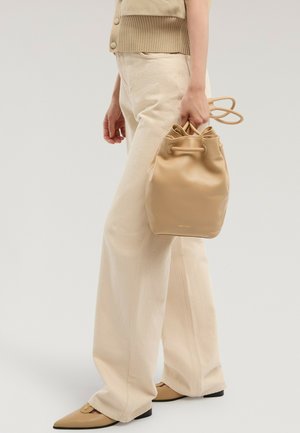 Osoba w beżowych szerokich spodniach i dopasowanych butach trzyma jasnobrązową skórzaną torbę typu bucket z ściągaczem za rączkę, widziana z boku.