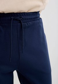 Camisa beige metida dentro de pantalones deportivos azul marino con cintura elástica y cordón atado en un lazo en la parte delantera.