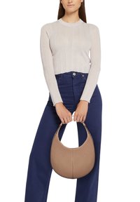 Borsa a tracolla in pelle beige con silhouette curva, tenuta da una persona che indossa un top a maniche lunghe a coste di colore rosa chiaro e jeans a gamba larga blu navy.