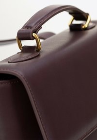 Ciemnobordowa skórzana torebka o sztywnej formie, z wyraźnie zaznaczonym uchwytem górnym zabezpieczonym złotym okuciem i starannym przeszyciem.