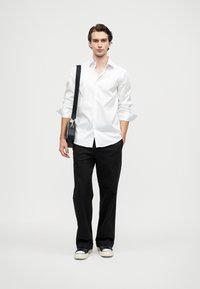 Vit skjorta med knappar, svarta byxor och marinblå sneakers. Modellen håller en svart axelremsväska. Slätt tyg och skräddarsydd passform, minimala detaljer.