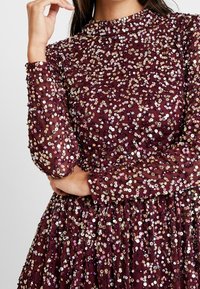 Kvinna i långärmad lila klänning täckt med iriserande paljetter med hög hals, som håller ena armen över midjan och handen nära ansiktet.