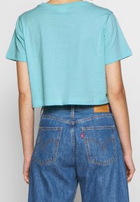 Persona que lleva una camiseta corta de color azul claro y jeans de mezclilla azul de cintura alta, vista desde la espalda contra un fondo liso.