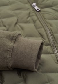Green quilted puffer jacket featuring a textured fabric, metal zipper, and a soft brown knit cuff detail.
