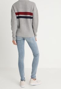 Szary sweter dzianinowy z poziomymi czerwonymi i granatowymi paskami na plecach, zestawiony z jasnoniebieskimi obcisłymi jeansami i beżowymi tenisówkami.