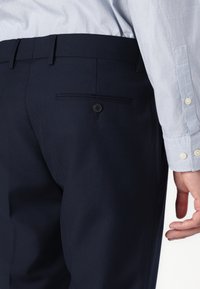 Navy blue tailored pants with a smooth texture, featuring a back pocket with a button closure and belt loops, paired with a light blue long-sleeve shirt.