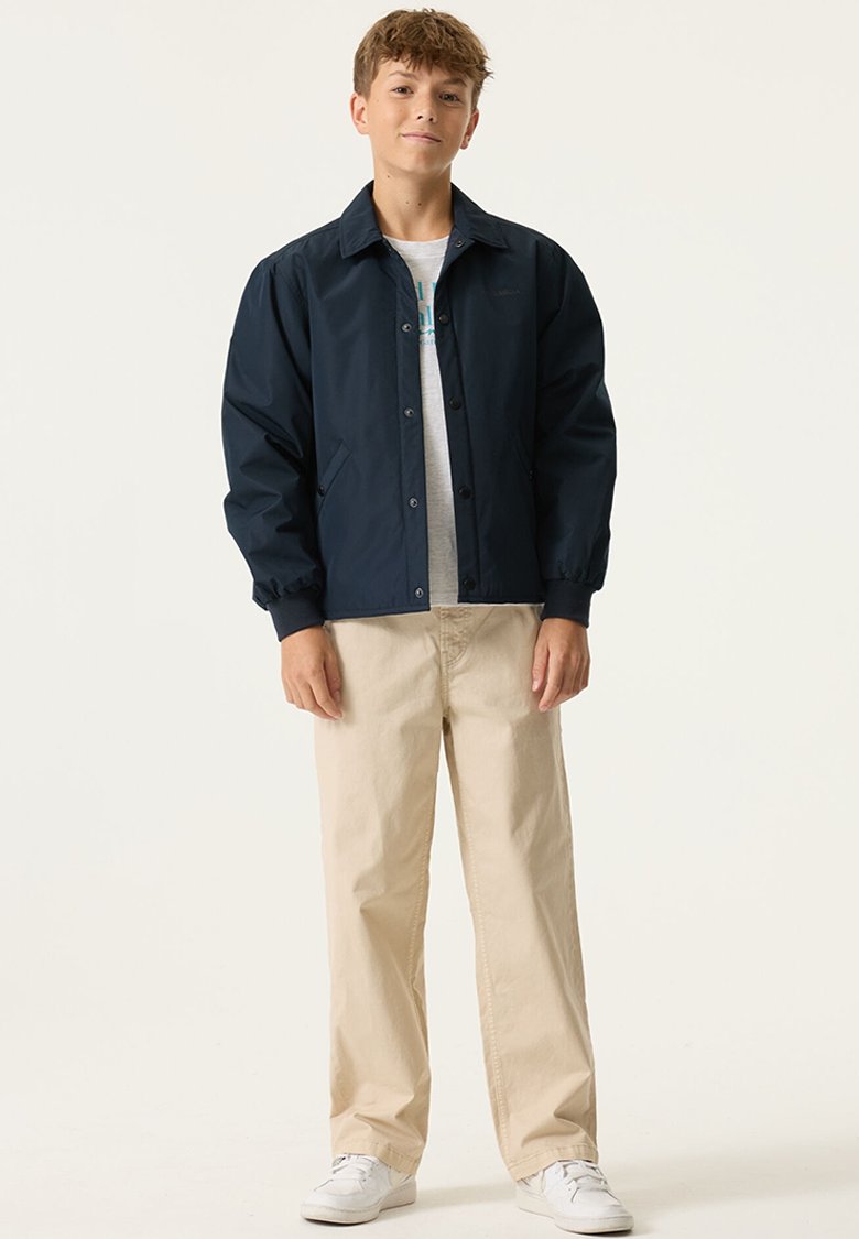 Chaqueta ligera azul marino con cuello de solapa, cierre de broches y bolsillos laterales, usada sobre una camiseta de cuello redondo y pantalones beige.