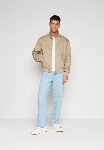 Veste bomber beige en tissu léger, avec une fermeture éclair à l'avant, des poignets côtelés et un petit logo vert sur la poitrine. Associée à un jean bleu clair.