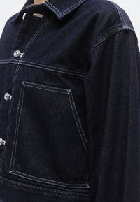 Sötétkék farmerdzseki strukturált gallérral, ezüst gombokkal, fehér varrással és nagy első zsebbel. Látható a texturált anyag részlete.