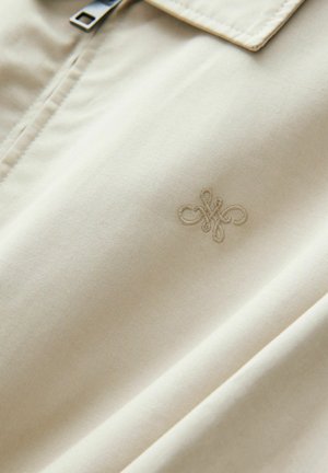 Ljust beige tyg med en slät yta; har en subtil broderad logotyp och en silverfärgad dragkedja vid kragen.